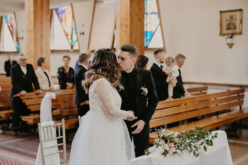 Para Młoda całuje się po ceremonii ślubnej w kościele, a goście stoją w ławkach Para Młoda całuje się po ceremonii ślubnej w kościele, a goście stoją w ławkach